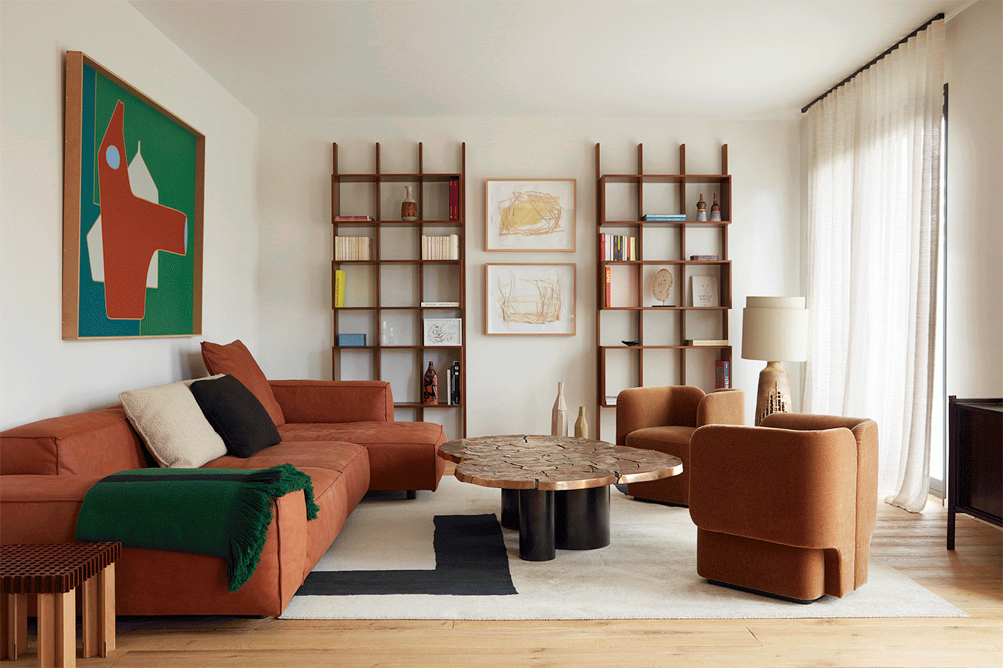 Neuilly Project by Sandra Benhamou - a red L shaped wall sofa with a centre table, two brown chairs, two wall shelf units and beige curtains.