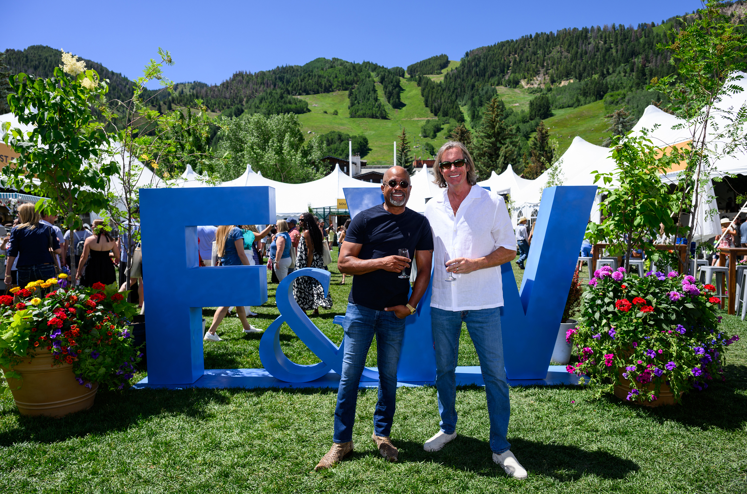Darius Rucker and Chris Radomski standing in front of a big blue sign saying 'F&amp;W' with white tents and green hills in the background
