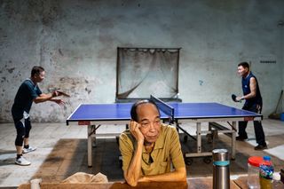 A man in a yellow shirt sits at a table, head resting on his hand, looking bored. Behind him, two others play ping pong in a dimly lit, worn-out room