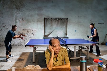 A man in a yellow shirt sits at a table, head resting on his hand, looking bored. Behind him, two others play ping pong in a dimly lit, worn-out room