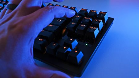 A hand hovers over the Razer Huntsman V3 Pro TKL 8KHz, glowing with amber backlighting. The scene is bathed in blue light, creating a tech-focused, futuristic atmosphere.
