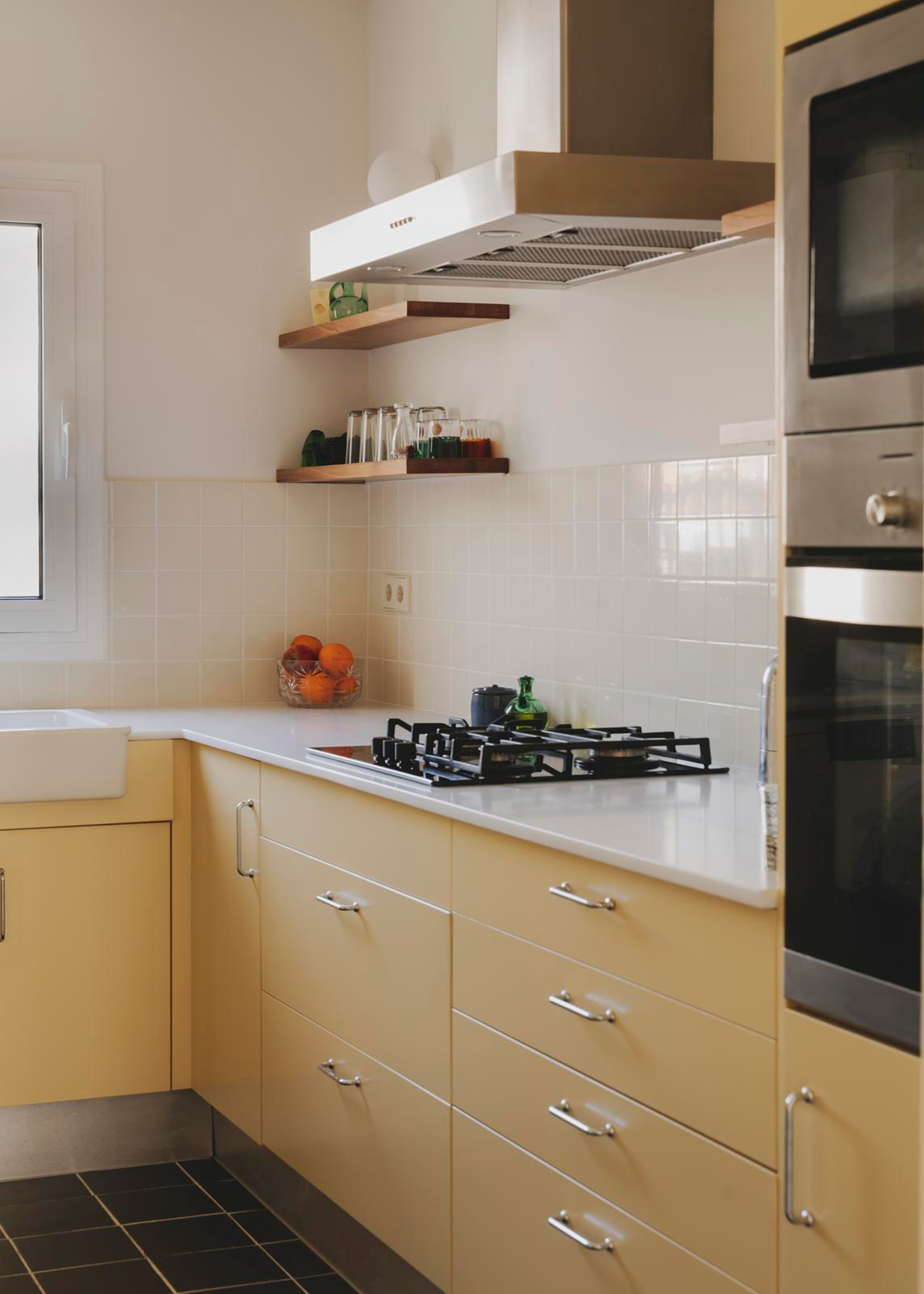 A small kitchen with a tile backsplash, a white stone countertop, butter yellow drawers and cabinets, floating wood shelves, a gas stove, and a black tile floor