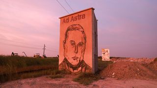 A portrait of a man is drawn on the side of a building next to a road at dusk.