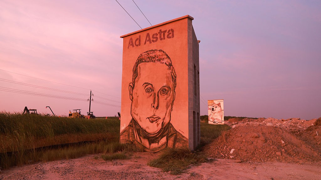 A portrait of a man is drawn on the side of a building next to a road at dusk.