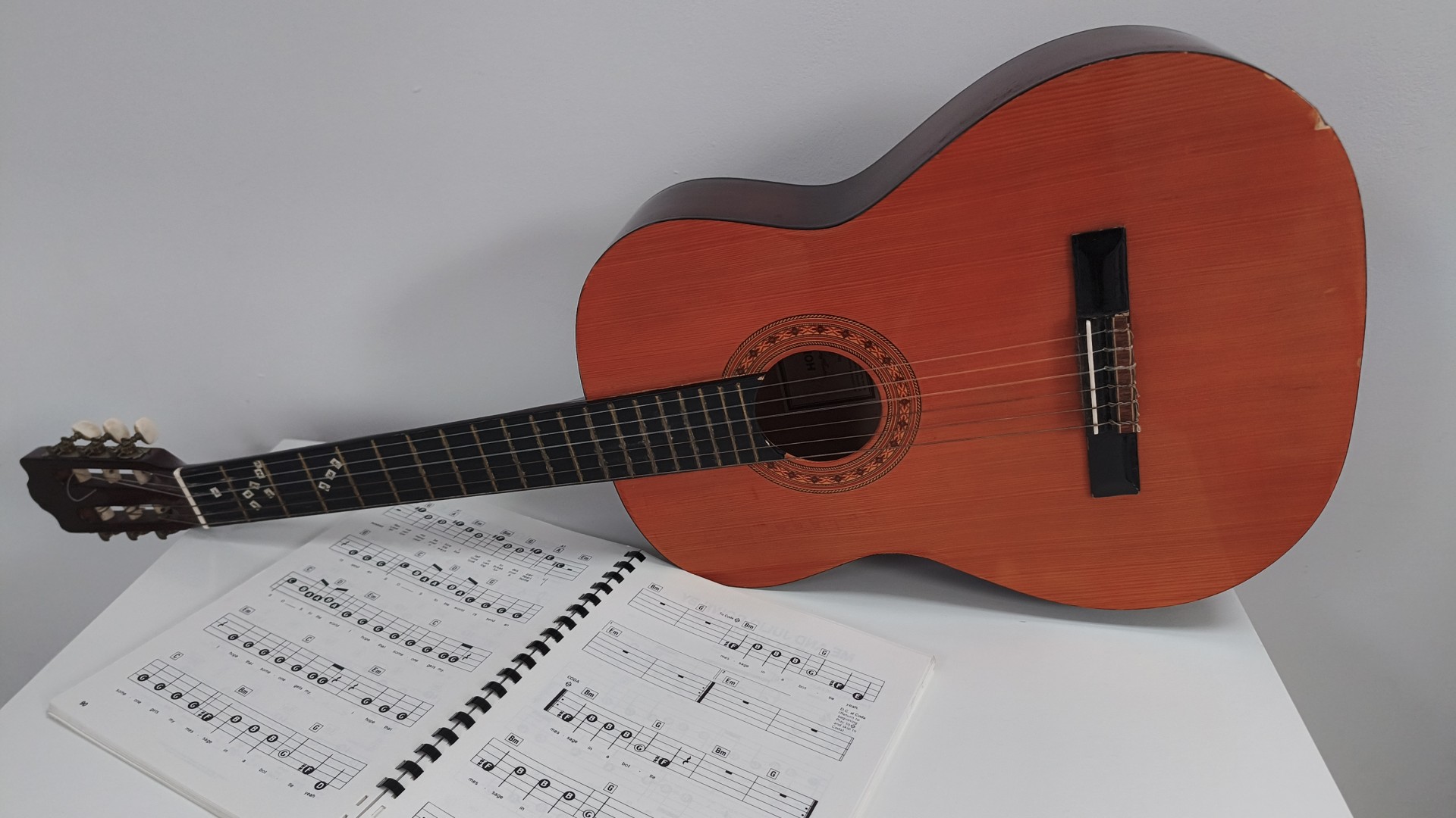 A classical acoustic guitar with nylon strings lies across an open spiral-bound book of sheet music on a white surface against a plain wall.