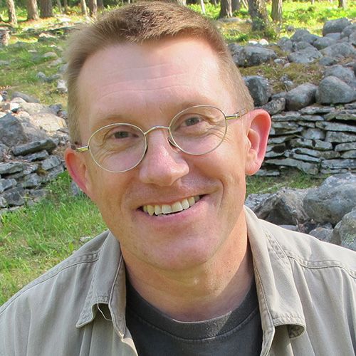 A man wearing a tan button down shirt and glasses stands in front of a series of stone structures.
