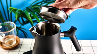 the fellow Corvo EKG kettle in matte black photographed against a blue background with a glass mug and black and white table