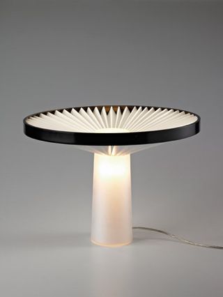A table lamp designed with a narrow illuminated stem and a circular fan-like lampshade with a black rim.
