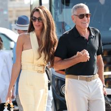 Amal and George Clooney walk outside in Venice. Amal wears a butter yellow halter-neck dress with white slingback heels. George wears a black polo top with white trousers and brown suede loafers.