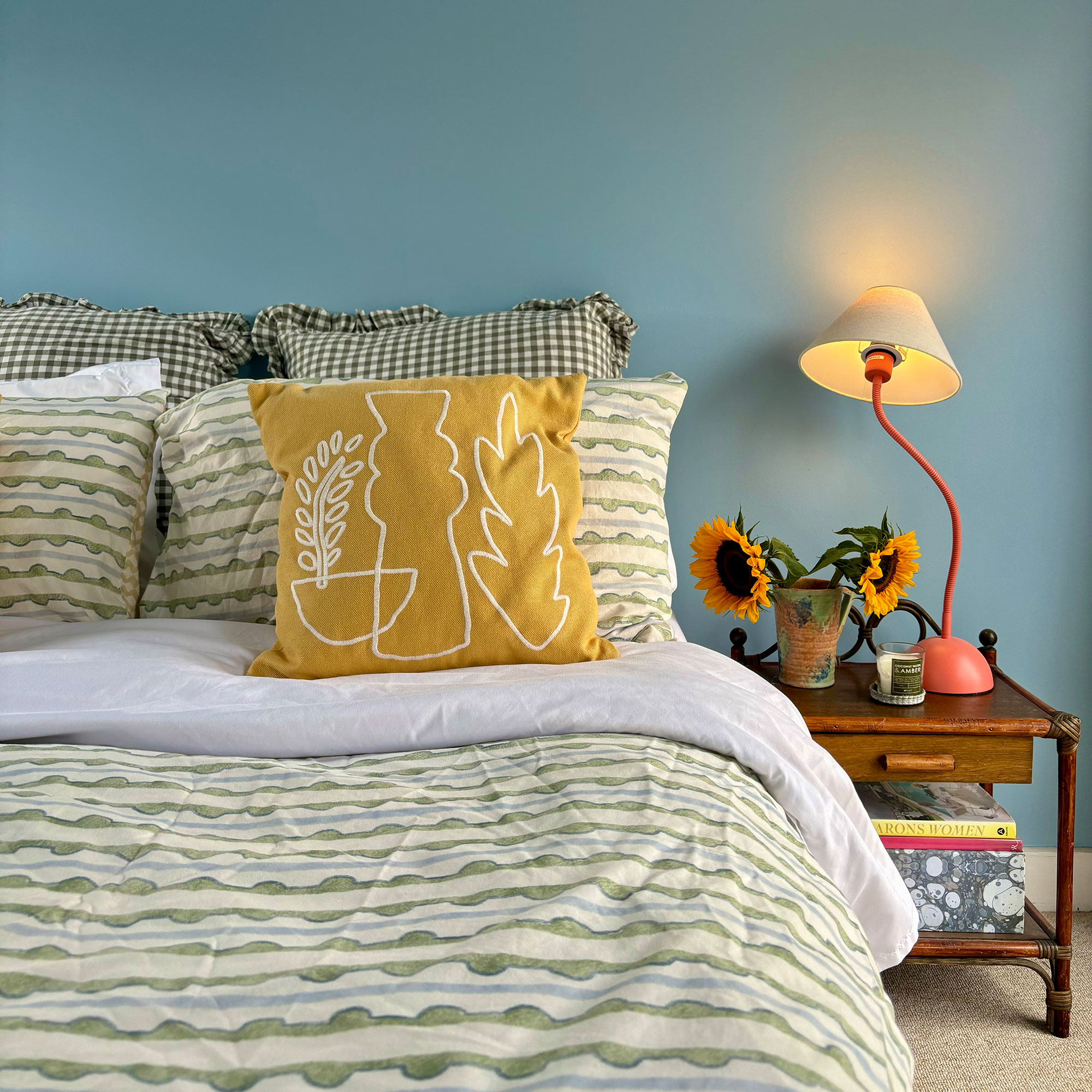 Blue bedroom with green and cream patterned bedsheets, pink wavy table light and vase of sunflowers