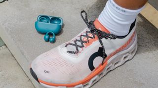 A teal pair of JLab Flex Open Clip earbuds and carry case on a concrete surface next to a foot in a white running shoe.