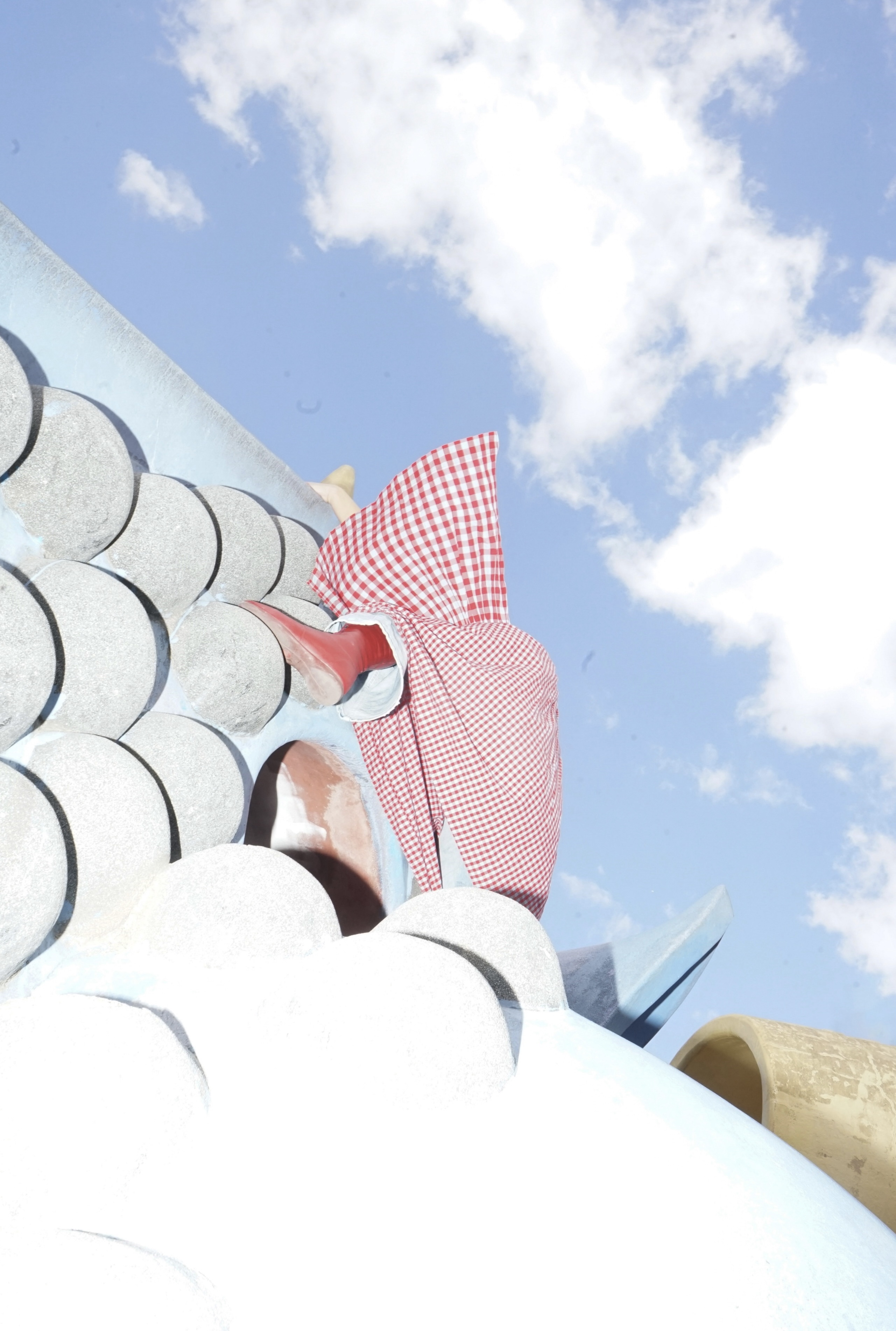 A person wearing a red and white checkered garment and a red boot climbs up a light-blue, scale-patterned playground structure under a cloudy blue sky.