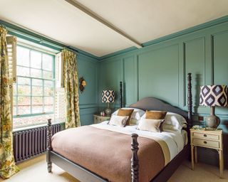 Main bedroom with blue green wood paneling and a double bed, floral yellow and green curtains, twin bedside tables and table lamps with a geometric print shade, black wooden bed frame, white and pale pink bedding and scatter cushions, cream carpet