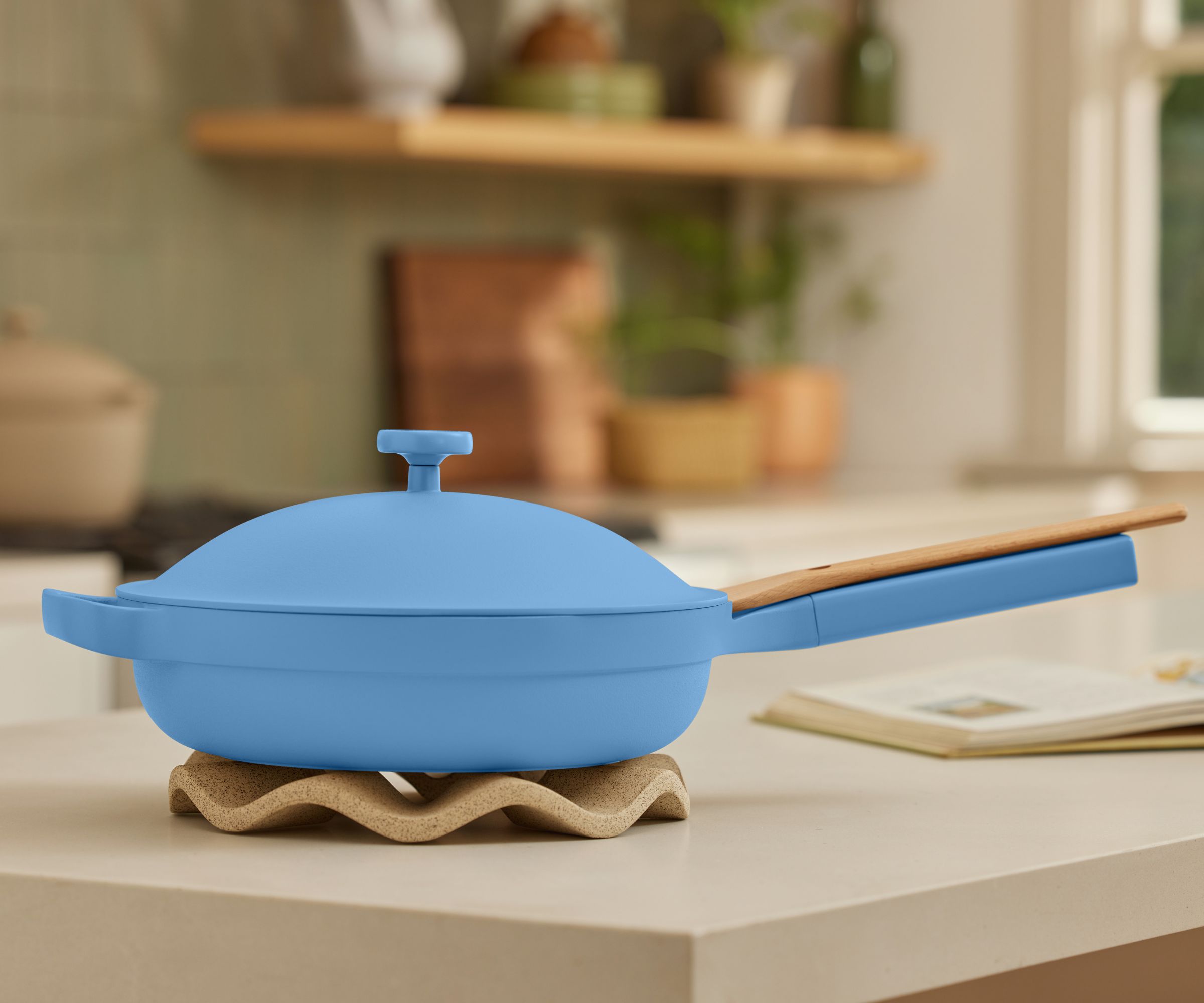 A blue pan with a long handle on one side and a carry handle opposite. A matching domed lid sits on top. The pan sits on a wavy trivet on a white quartz countertop, and there is an open cookbook out of focus to it's right.