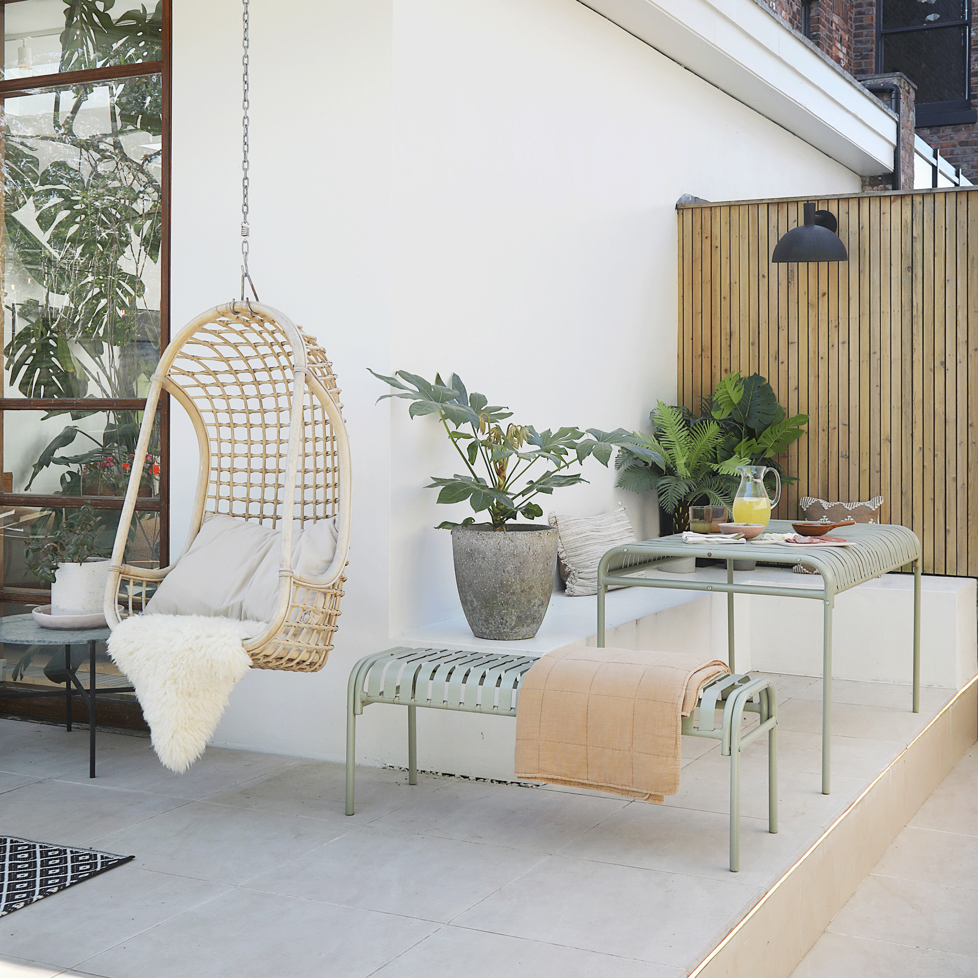 a patio area with a metal dining table and bench with a hanging wicker egg chair