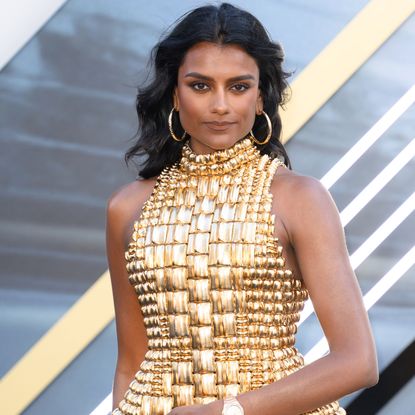 Simone Ashley wearing a gold halter neck top. Her hair is curled and she is also wearing gold hoop earrings.