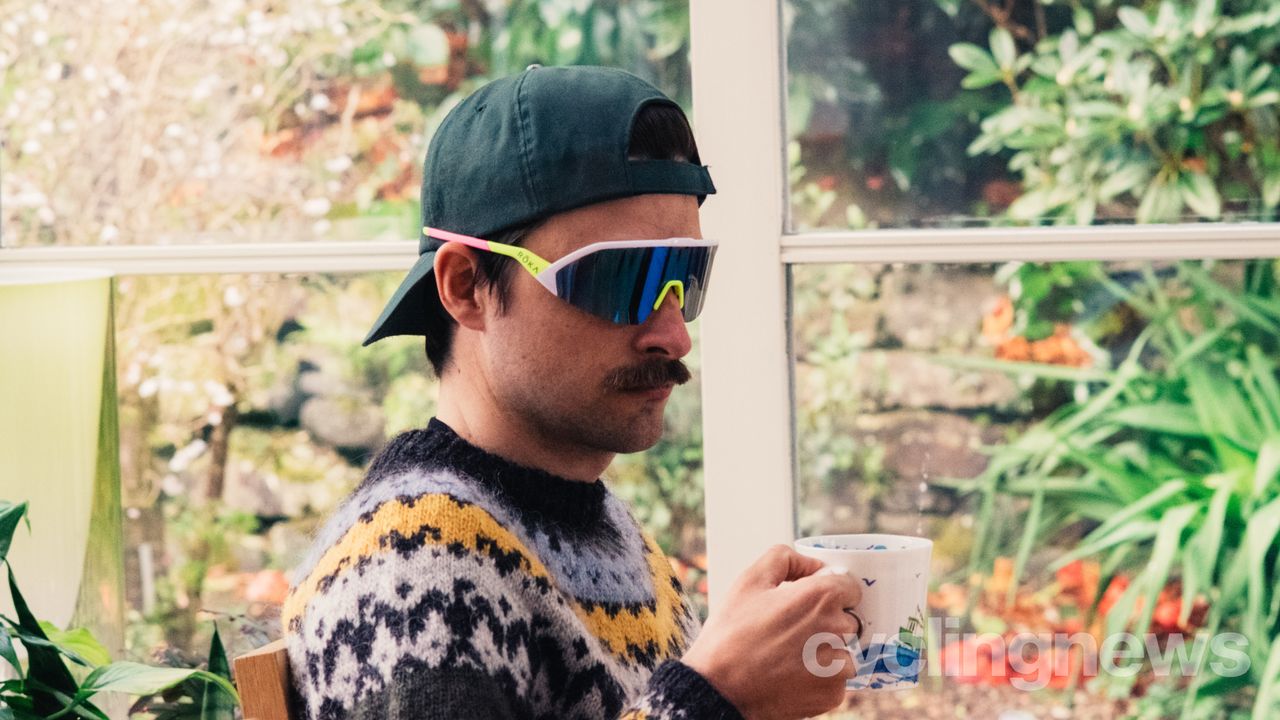 A white man in a backwards cap and a colourful jumper holds a mug of tea while wearing brightly coloured sunglasses