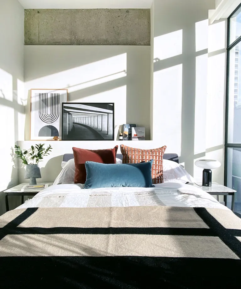 An apartment bedroom with scatter cushions in red ochre and teal, and artwork leaning on a shelf