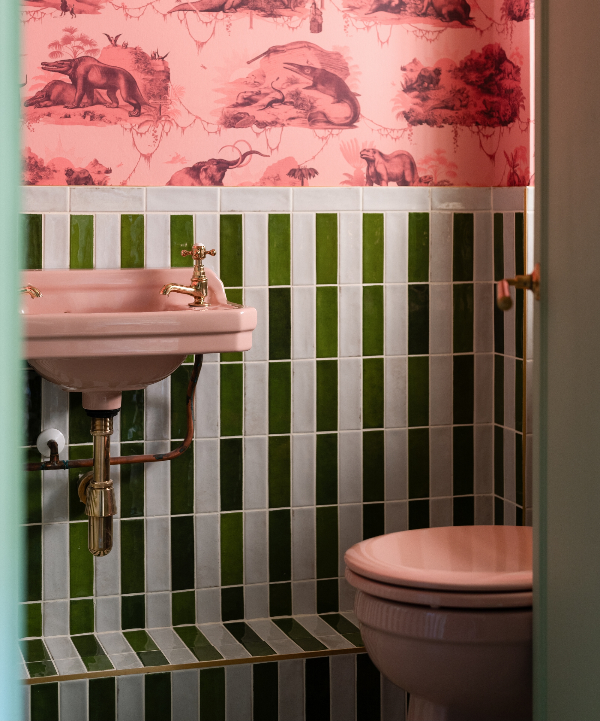 Pink sink and toilet against green striped tiles, brass taps, playful pink animal print wallpaper above.