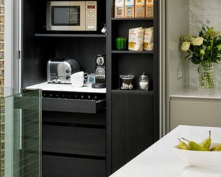 A modern kitchen pantry with microwave, coffee maker and pears in bowl on white island