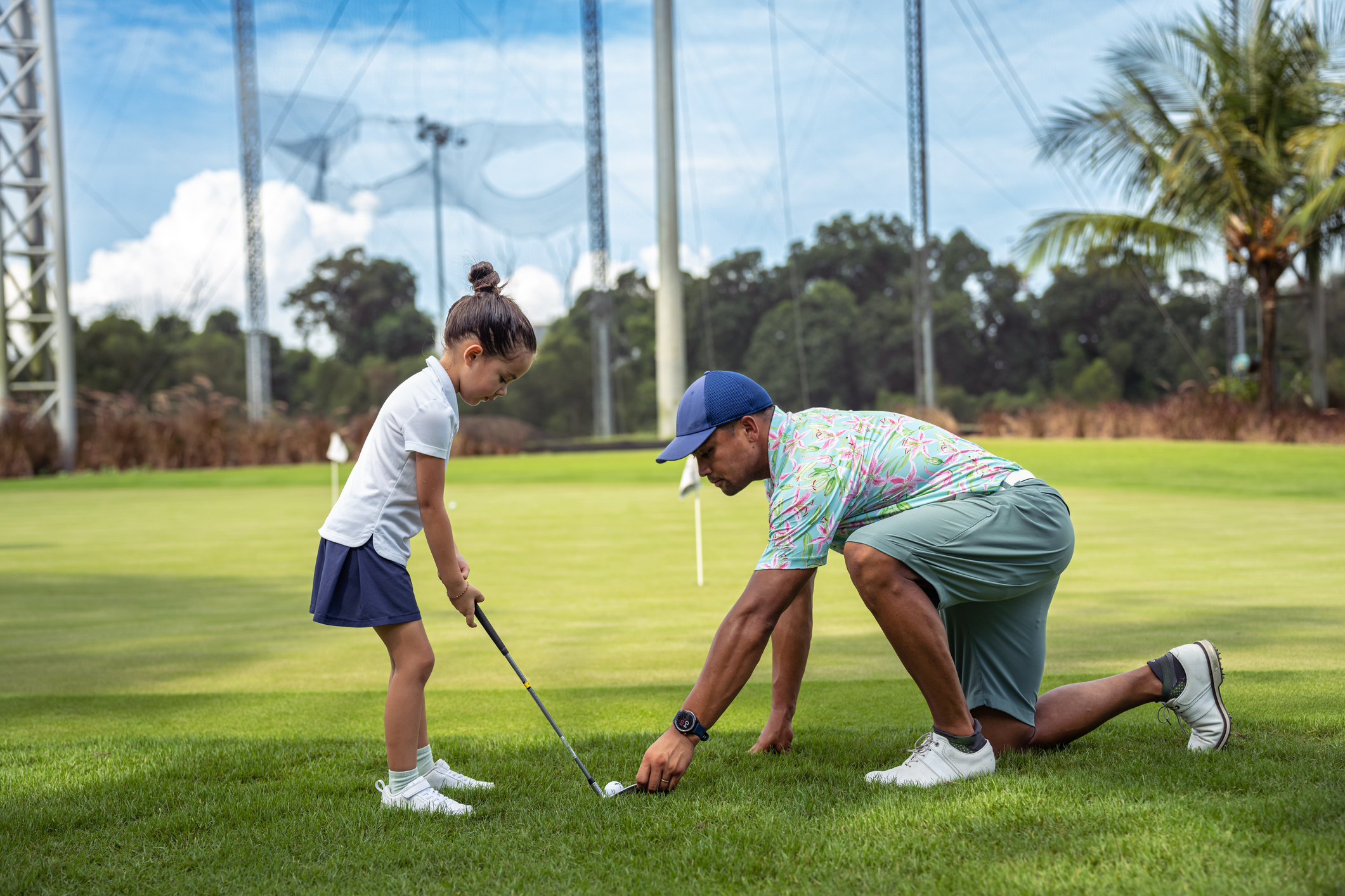 A father kneels on the golf course, repositioning a golf ball as he instructs his kindergarten aged daughter on how to putt.