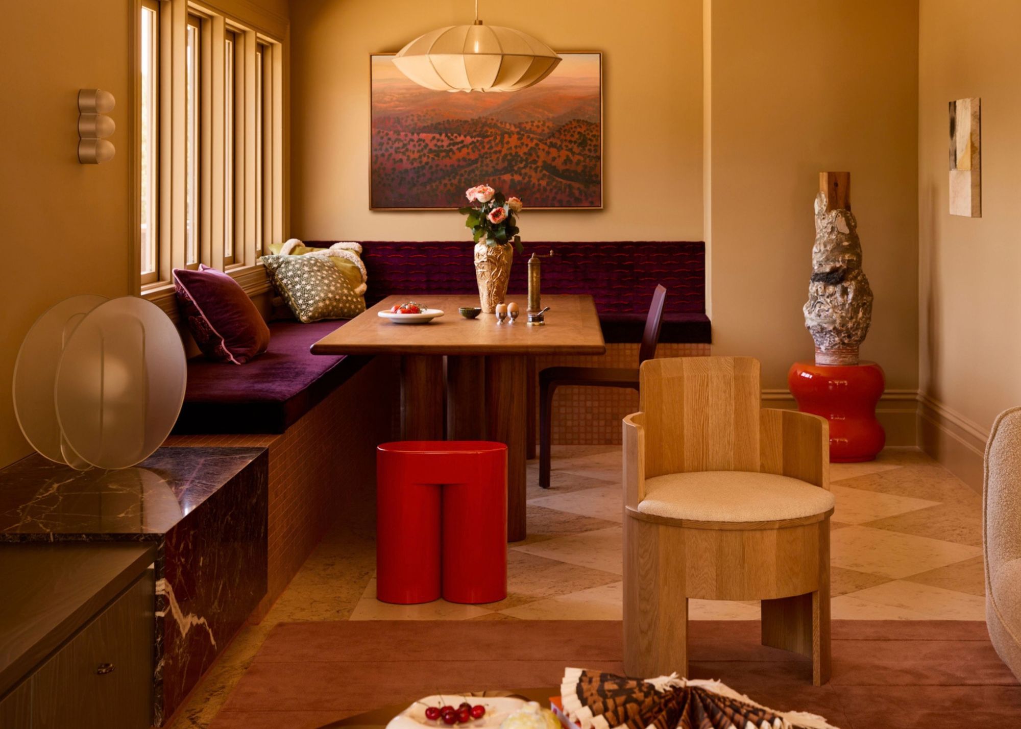 A modern open plan space with a living and dining area with a banquette with purple seating on a wooden bench with a large pendant light above the table and two bold red stools