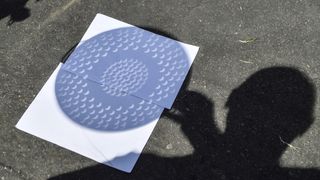image of a shadow of a kitchen colander on white pieces of paper showing a partially eclipsed sun in the holes.