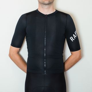 A white man in a black cycling jersey and black cycling shorts stands against a white wall