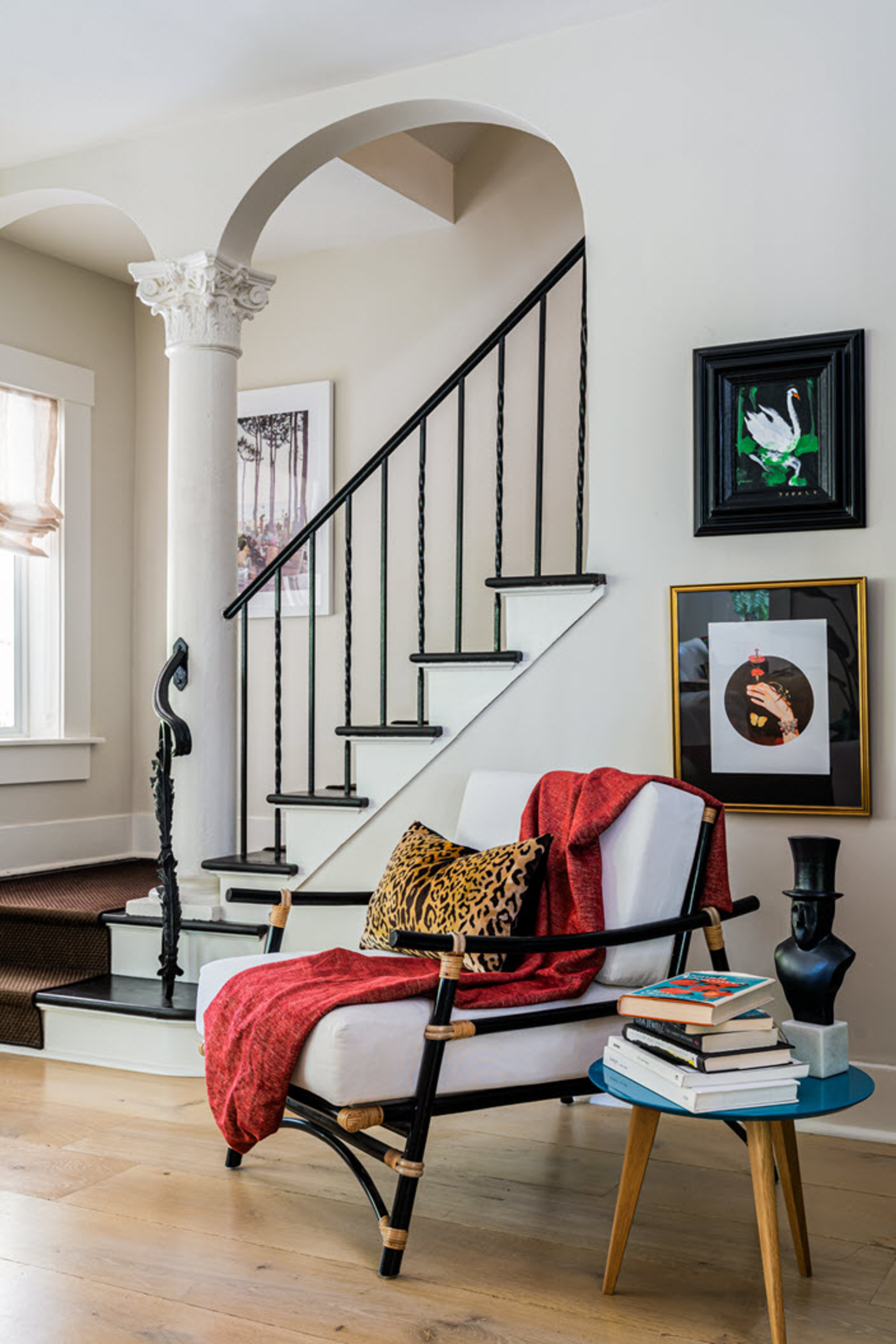 an ornate staircase with columns and black tread in an otherwise modern space with timber floorboards, artwork on the wall, and an armchair draped with a red throw and leopard cushion next to a side table stacked with books