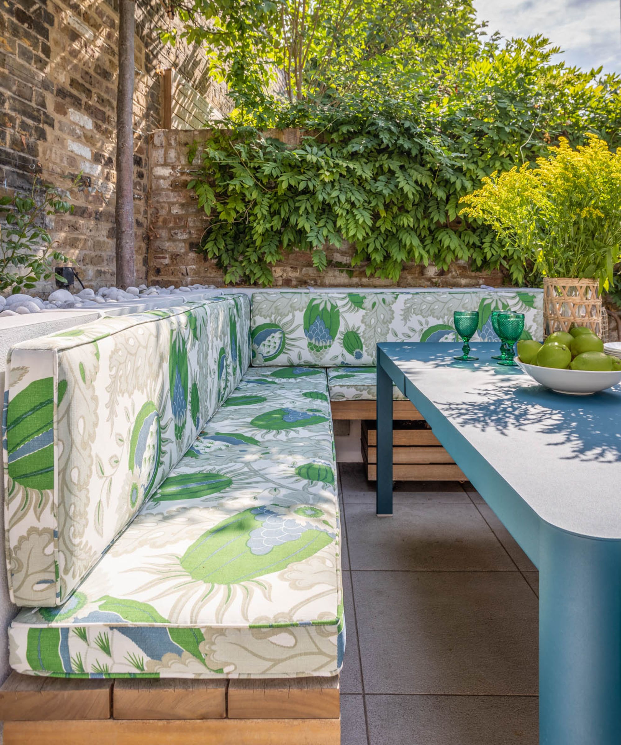 a walled garden with a wooden banquette seat and floral upholstered bench cushions iwth blue dining table