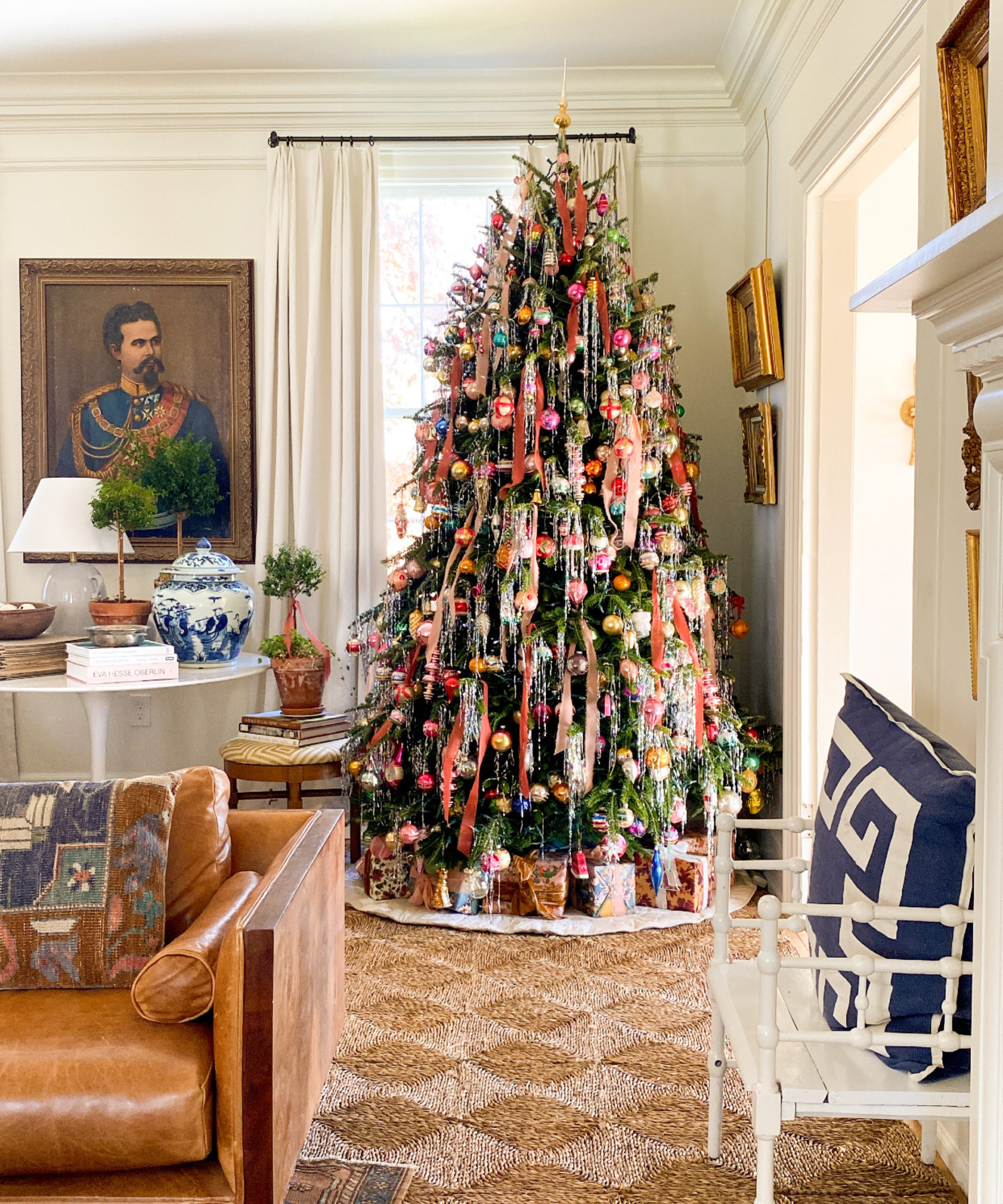 a living room with a large Christmas tree decorated with ornaments, bows, and tinsel. A jute rug rests on the floor, chairs, a couch, and a table are also in the room