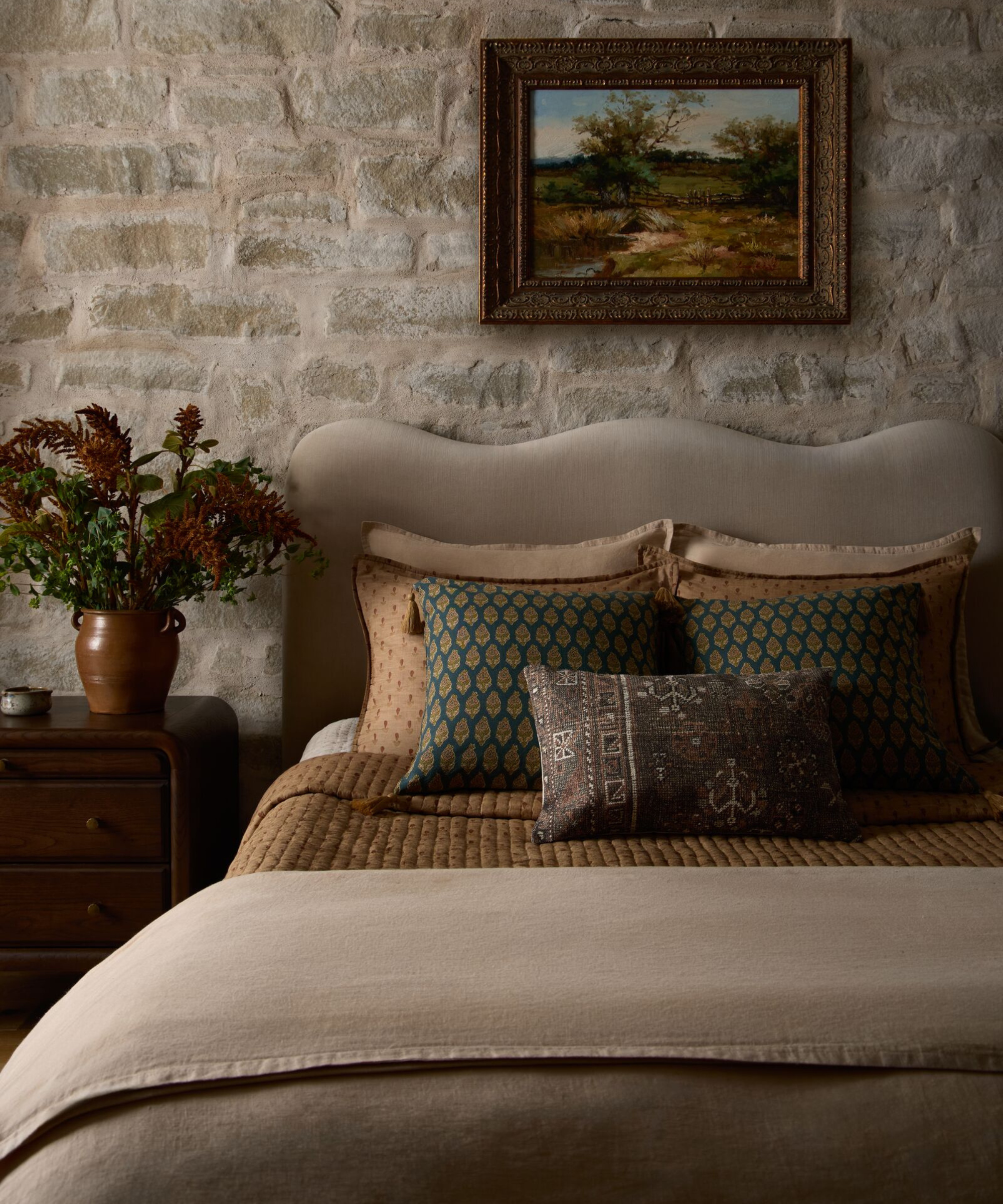 a cozy bed with many pillows, a taupe bedspread, and a stone wall