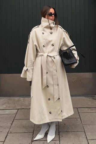 Spring trench coats shown in a photo of a standing outside in Stockholm wearing black sunglasses, with a funnel-neck trench coat, black tote bag, gray socks, and white slingback heels