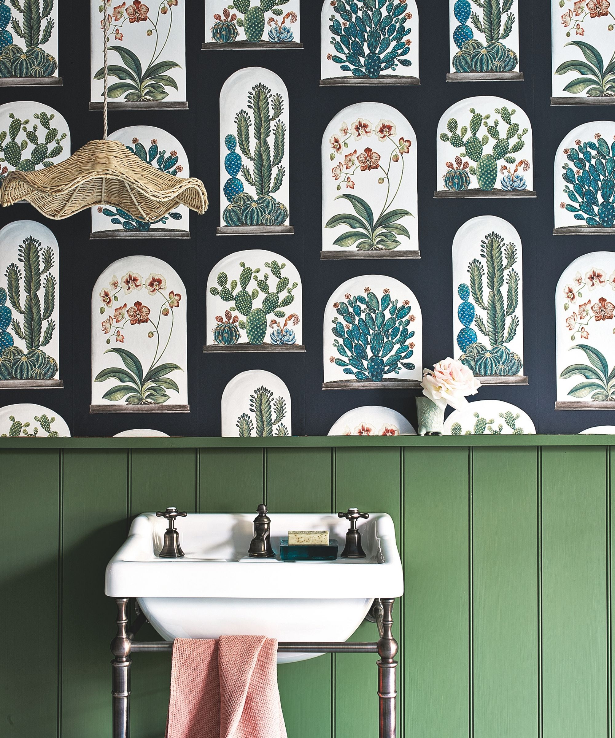A white porcelain sink on a metal stand with matching faucets, with a green painted wall panelling behind. Above the panelling is a botanical print black wallpaper with cacti in terrariums. There is also a hanging lamp with a rattan shade.