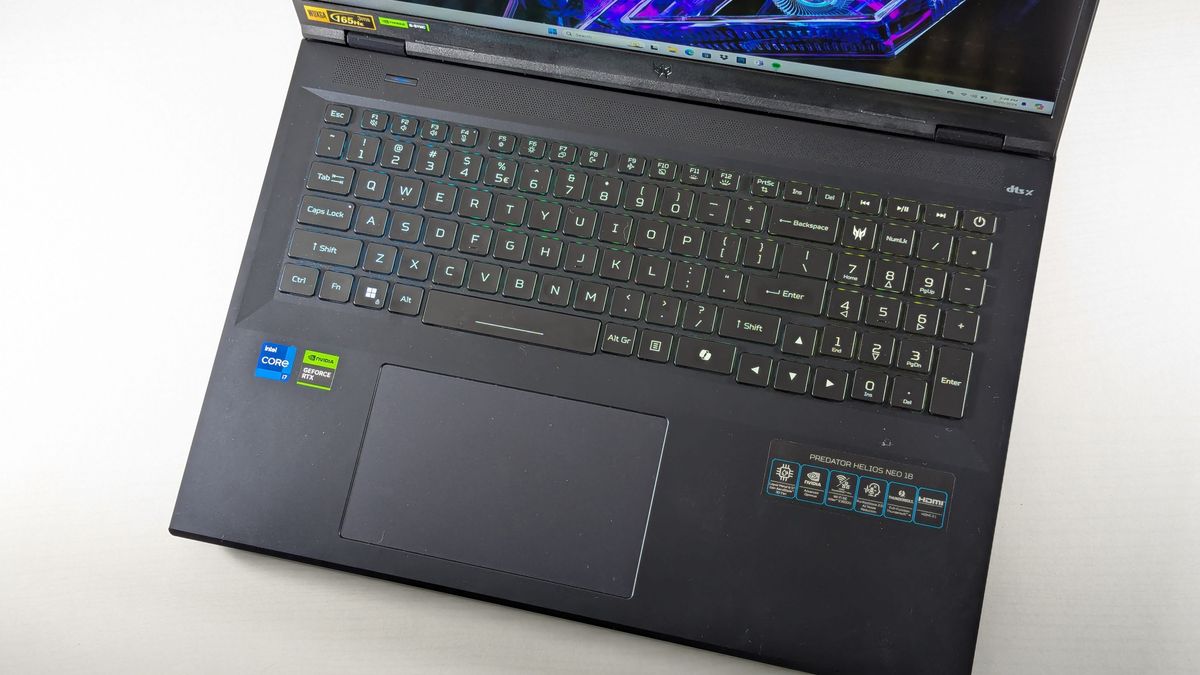 An angled view of the backlit keyboard and trackpad on the open and powered on Acer Predator Helios Neo 18 on a light gray background.