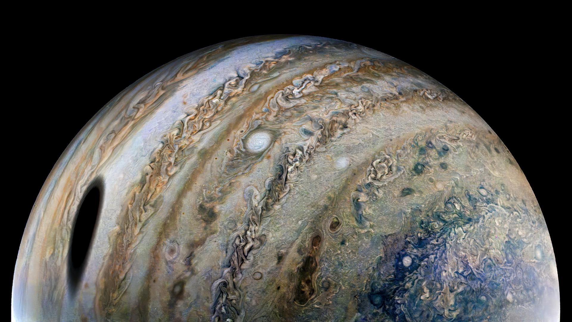 View of one-half of Jupiter. Swirling rings of gold, brown, green, and blue dust and gas clouds wrap vertically around the sphere of the planet. A large dark spot is seen toward the bottom left and smaller spots of white dot the surface, breaking up the rings of swirling clouds and dust. 