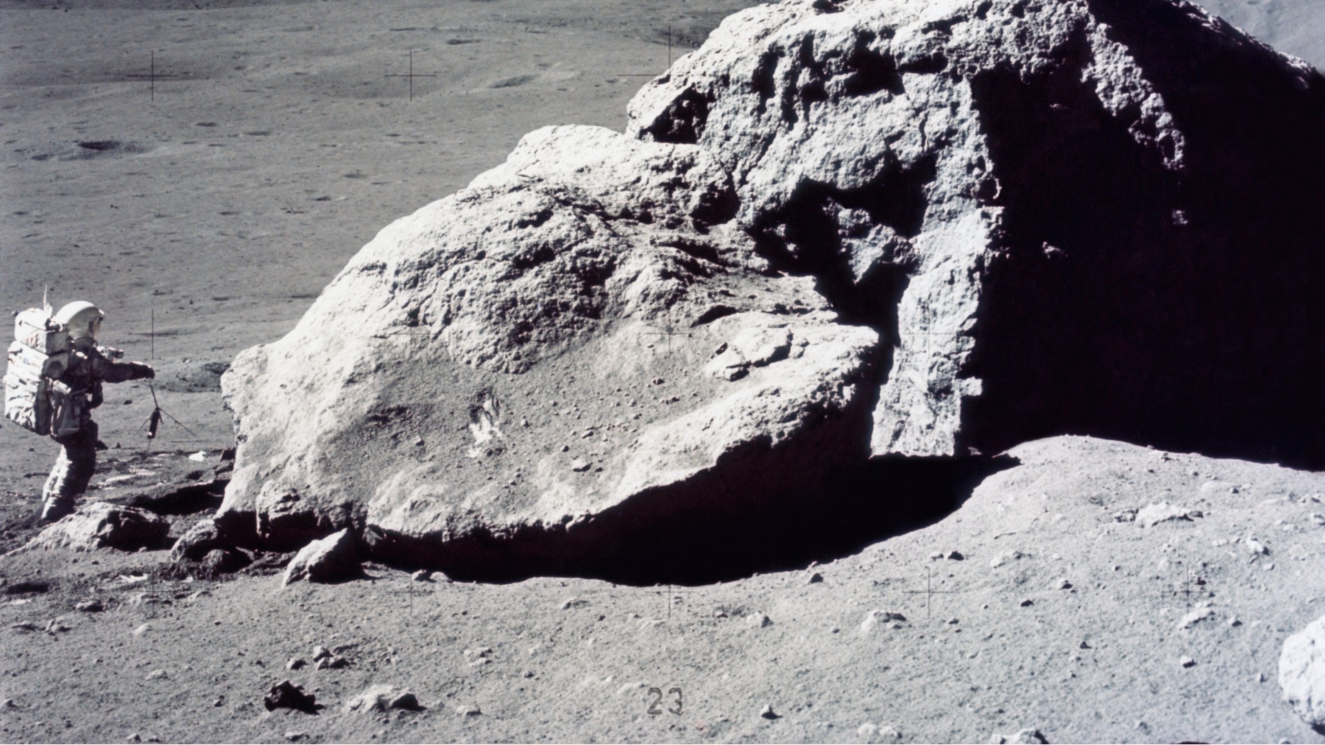 يقف رائد فضاء يرتدي بدلة فضائية ضخمة بجوار صخرة كبيرة على سطح رمادي محبب