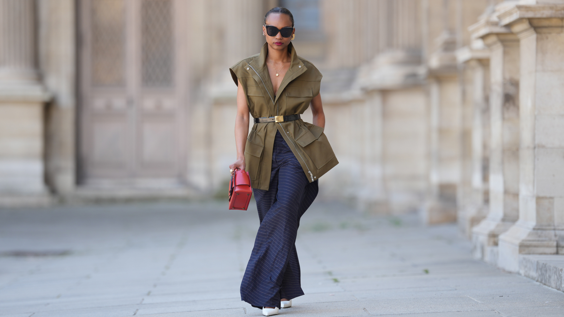 a woman wears a long blue skirt, a khaki top, and a red bag while she walks through a European city