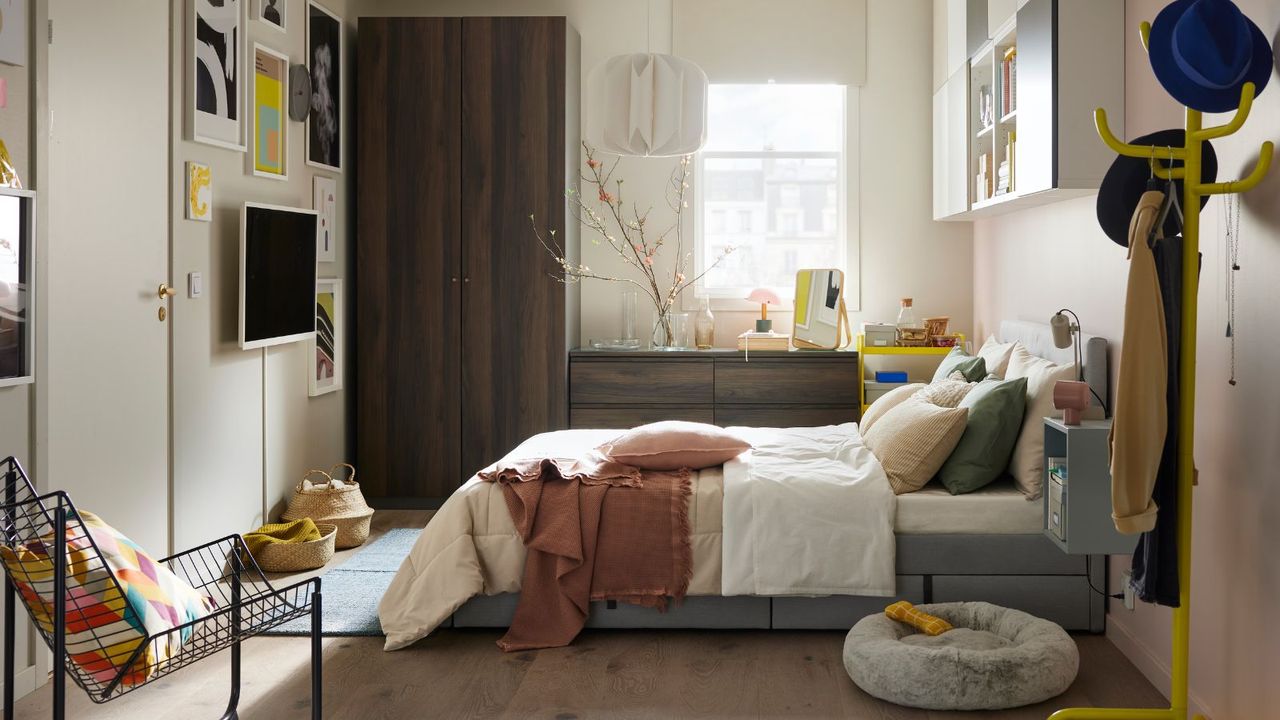 Beige and white bedding on a bed in front of a window, surrounded by a chest of drawers, a chair, and a hat stand. 