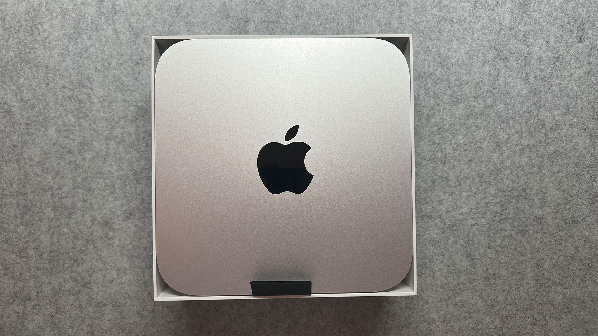 The Mac Mini Pro M2 is mini pc. It's a thin, square shape with rounded corners (20 x 20 x 3.5 cm). It's silver in color and has the Apple logo in the center of it (a black apple with a bite taken out of it). It is snuggly inside a square box.