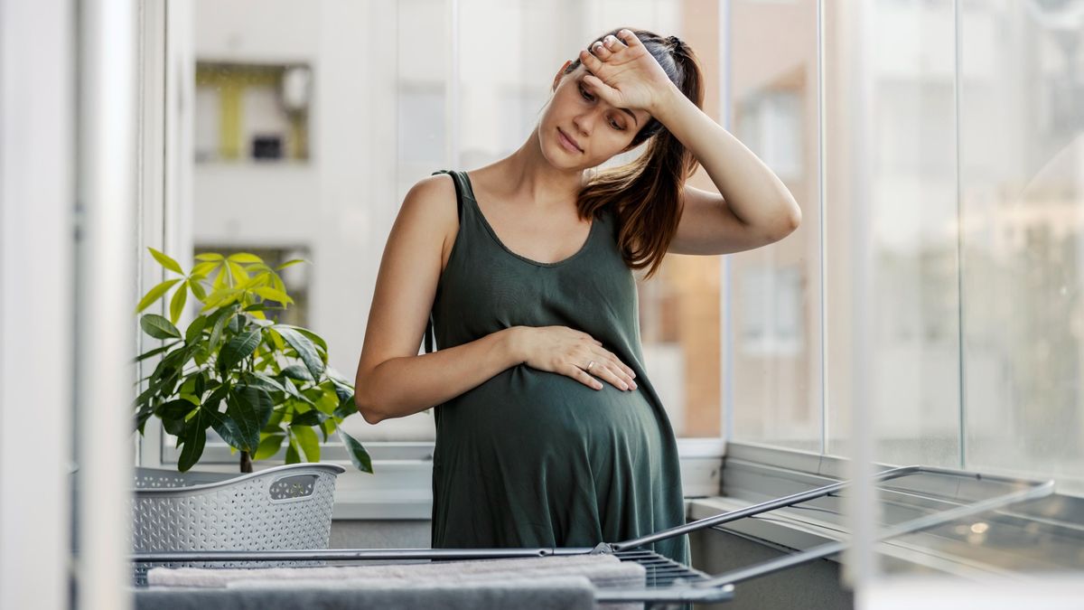 A pregnant young woman is shown standing by a window. She has her hand over her belly and the other hand wiping sweat across her forehead.