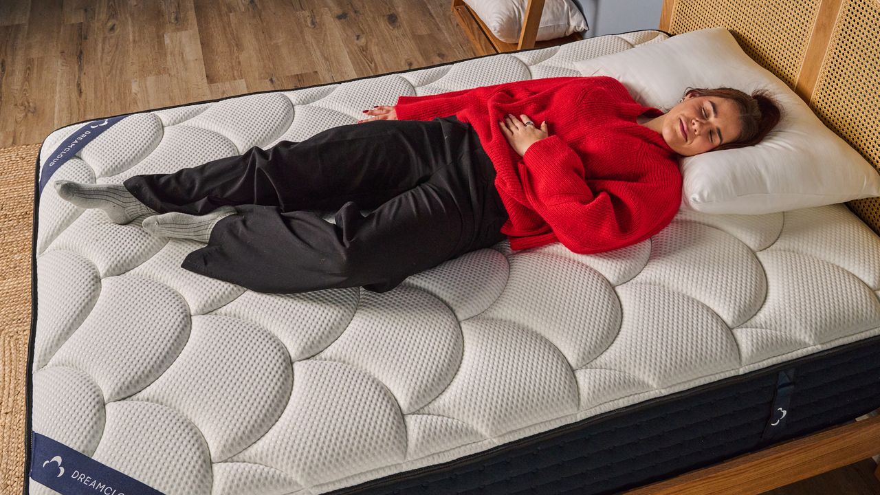 The image shows a woman wearing a red jumper and black trousers lying on top of the DreamCloud Hybrid Mattress
