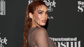 Megan Fox attends the Sports Illustrated Swimsuit issue party in 2023. She is standing on the red carpet and staring at the camera in a long sleeve dress. Her hair is wet.