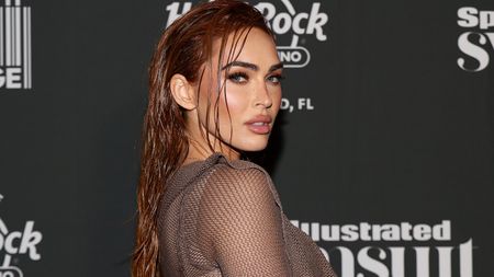 Megan Fox attends the Sports Illustrated Swimsuit issue party in 2023. She is standing on the red carpet and staring at the camera in a long sleeve dress. Her hair is wet.