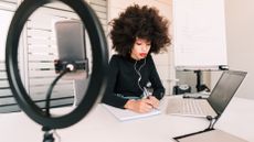 Woman working on a laptop wiith a camera and ring light pointed at her