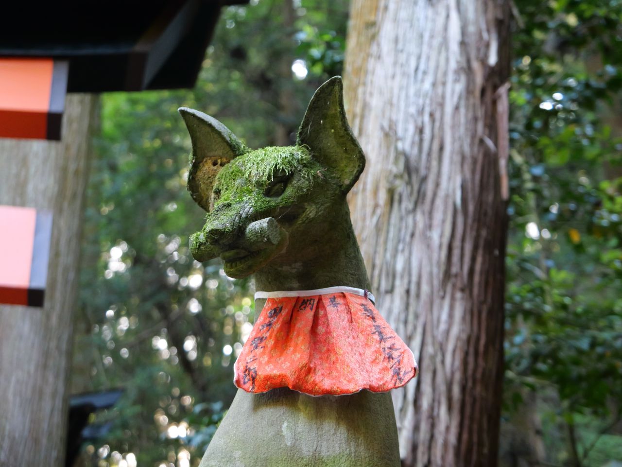Moss-covered fox statue wearing a red bib at a shrine.