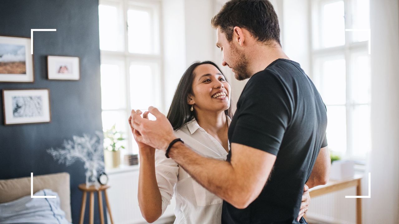 Man and woman smiling and dancing together in the living room of their home with couch and table in the background, representing is it important to have sex