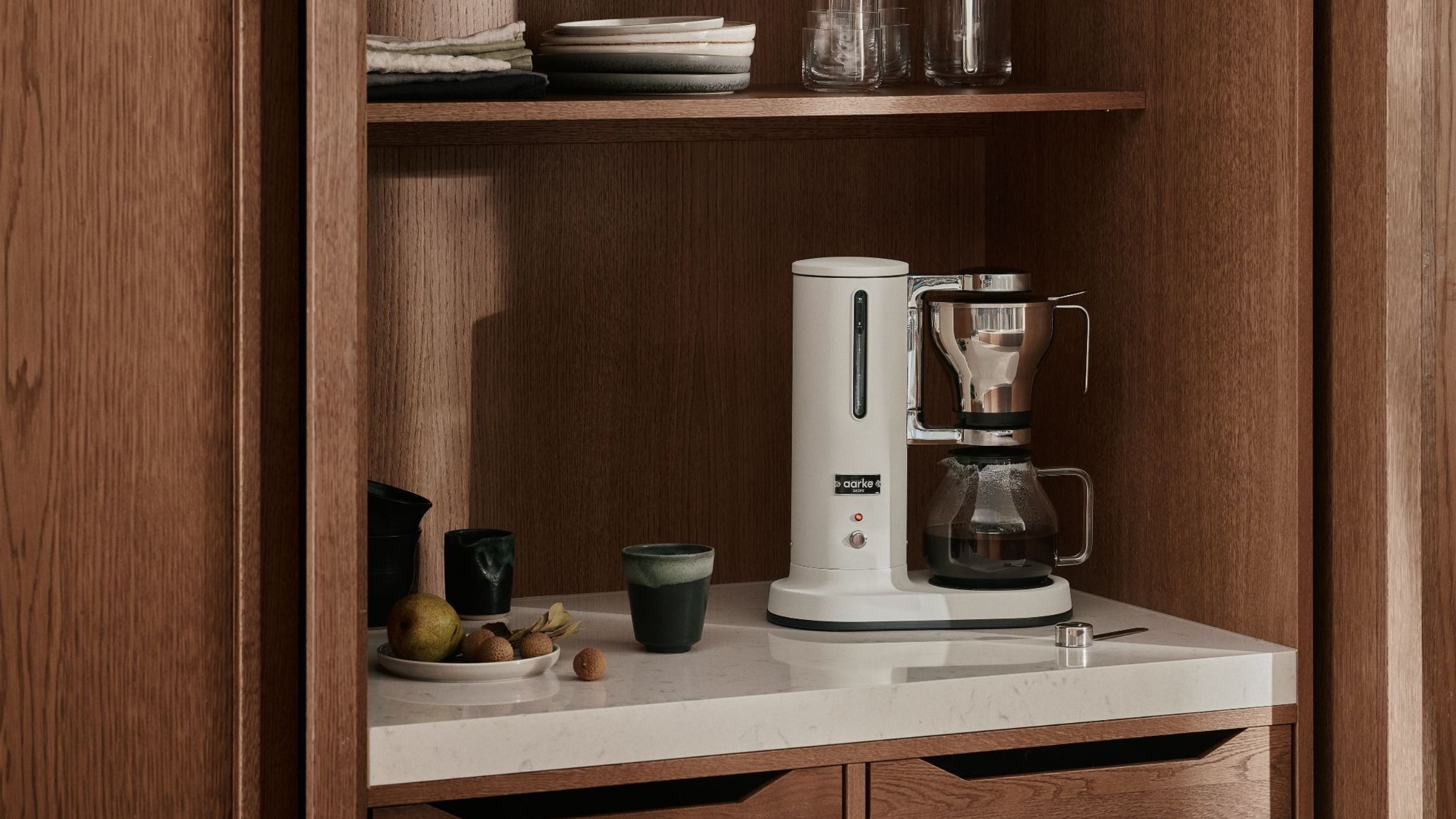 A white Aakre drip coffee maker tucked into a dark wood appliance garage with white counters. 