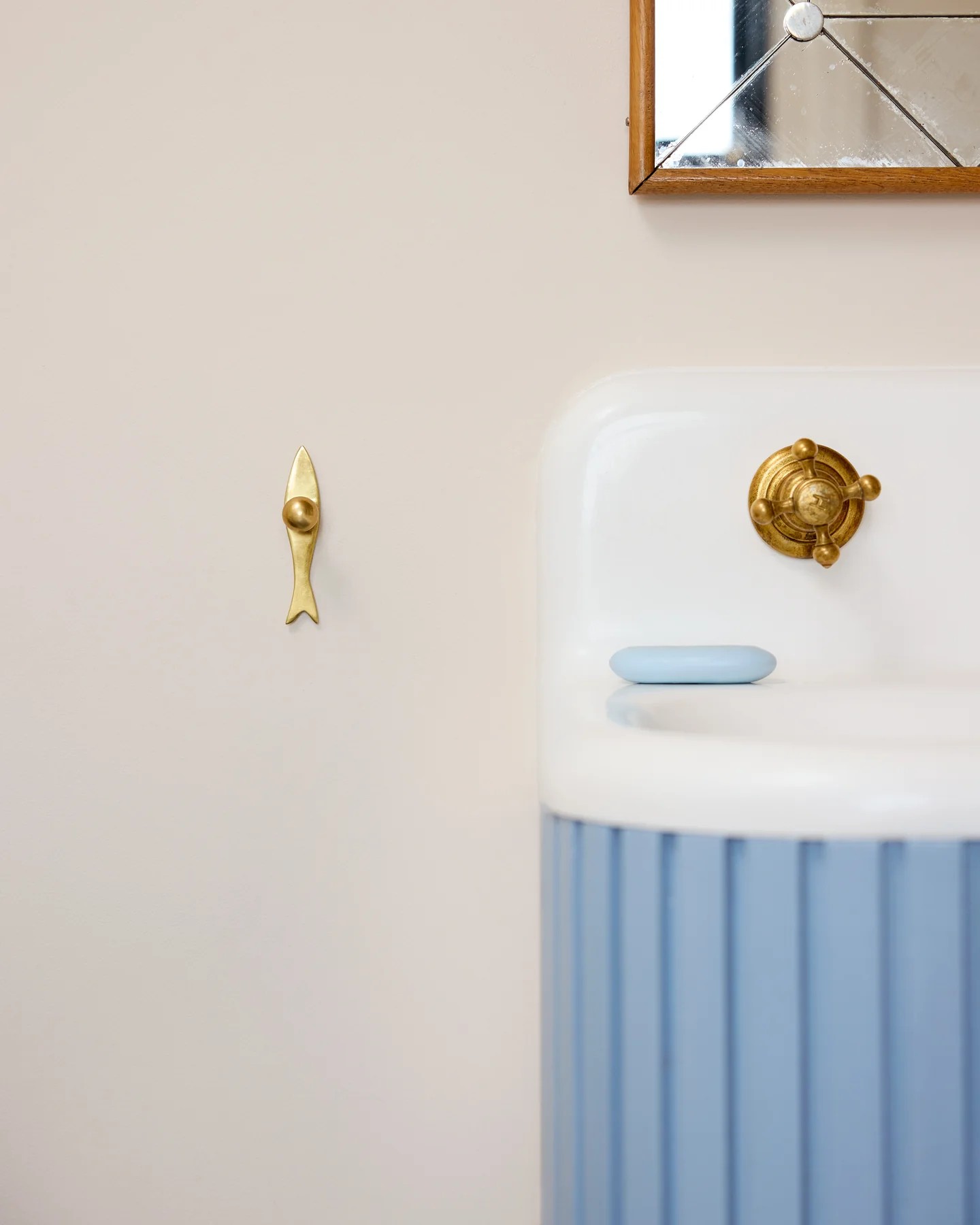 Image of a brass, fish-shaped hook hanging on an off-white bathroom wall. There is a light blue and white sink with a blue bar of soap on it in view.