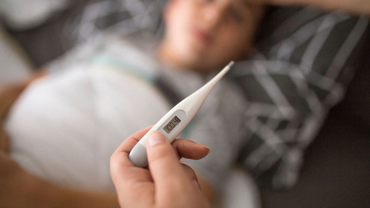 close up of an adult&#039;s hand holding a thermometer that reads 100.4. A young boy can be seen in the background under a blanket, as if ill